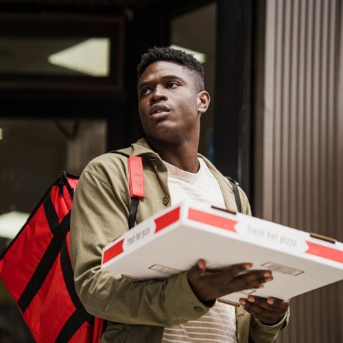 pexels-photo-13443803-13443803 A delivery person holds pizza boxes outdoors with a red backpack, ready for delivery.